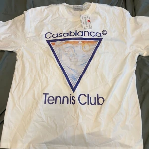 Vit Casablanca Tennis Club t-shirt - Casablanca t-shirt i vit bomull med stort färgglatt tryck på framsidan som visar en våg, sol och moln i en triangel. Snyggt 'Tennis Club'-print i blått under motivet. Klassisk passform och rund hals. Perfekt för dig som gillar sportig och trendig stil.
