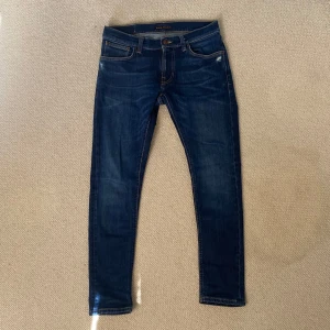 Mörkblå jeans från Nudie - Säljer ett par mörkblå skinny jeans från Nudie med klassisk femficksdesign och snygga kontrastsömmar. Jeansen har låg midja och är tillverkade i stretchig denim för en tight passform. Bakfickorna har Nudies ikoniska broderi. Köpt från Nudie butiken och i fint skick