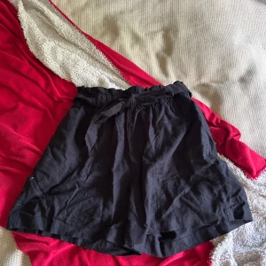 Svart kortkjol med knyt från Esmara - Svart kortkjol från Esmara med elastisk midja och snyggt knytband framtill. Kjolen har en lätt och luftig känsla, perfekt för varma dagar. Stilren design som är enkel att matcha med olika toppar och sneakers.