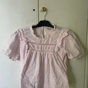 Ljusrosa blus med volang från Gina Tricot - Supersöt ljusrosa blus från Gina Tricot med korta puffärmar och med knapp i nacken. Använd 1 gång och är i ett mycket bra skick! Kan vara någon skrynklig men det går lätt att fixa genom att stryka den lite💞