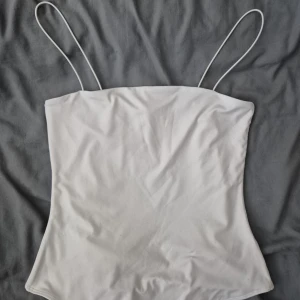 Vit bodysuit med tunna axelband H&M - Säljer en stilren vit bodysuit från H&M med tunna axelband och rak halsringning. Bodyn har en clean look och är tillverkad i mjukt stretchigt material som sitter snyggt på kroppen. Perfekt att styla med jeans eller kjol för en enkel och fräsch vibe.