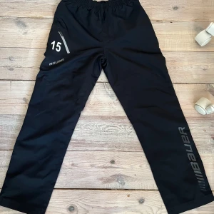 NYA Bauer överdragsbyxor  - Svarta Bauer överdragsbyxor i juniorstorlek, helt nya och aldrig använda. Nummer 15 på höger lår och Bauer-logga längs vänster ben. Byxorna har dragkedja vid ficka samt resår i midjan för bekväm passform.  Perfekta för hockey, uppvärmning, träning eller som bekväma byxor till vardags. 