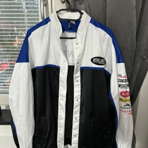 Racinginspirerad vindjacka H&M Divided XS - Snygg racinginspirerad vindjacka från H&M Divided i färgerna svart, vit och blå. Jackan har tryck med text och loggor på bröstet, ärmen och ryggen, samt knäppning framtill. Perfekt för dig som gillar streetstyle och sportig vibe.