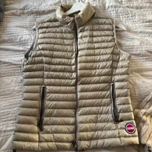 Beige dunväst från Colmar - Snygg beige dunväst från Colmar med quiltad design och hög krage. Västen har två dragkedjeförsedda fickor och en tydlig Colmar-logga framtill. Perfekt att slänga över en hoodie eller tröja för extra värme och stil.