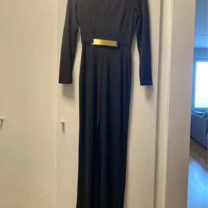 Svart långklänning med guldigt bälte - Supersnygg svart långklänning med lång ärm och ett stilrent guldigt bälte i midjan som ger en cool touch. Klänningen har en smickrande, figurnära passform och är gjord i ett mjukt, följsamt material. Perfekt för dig som gillar minimalistisk och elegant stil.