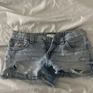 Ljusblå jeansshorts från Gina Tricot - Snygga ljusblå jeansshorts från Gina Tricot med slitna detaljer och råa kanter. Shortsen har fem fickor, dragkedja och knapp framtill samt broderade bakfickor med paisleymönster. Perfekta för sommarens chill dagar.