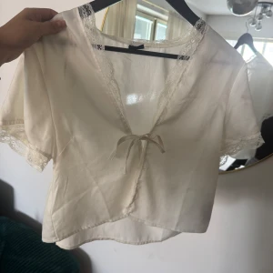 Vit omlottopp med spets från Gina Tricot - Supersöt vit omlottopp från Gina Tricot i satinliknande material. Toppen har korta ärmar, spetsdetaljer längs kanterna och en liten rosett framtill. Perfekt för dig som gillar romantiska och feminina vibes.