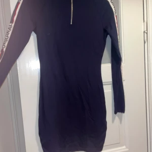 Mörkblå sportig klänning med dragkedja - Sportig mörkblå kortklänning med långa ärmar och vit/röd rand längs ärmarna där det står 'ORIGINAL'. Halvlång dragkedja framtill med rund metallring som detalj. Skön passform och perfekt för en avslappnad streetstyle-look.