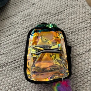 Holografisk mini-ryggsäck med regnbågspuff - Säljer en liten ryggsäck i holografiskt syntetmaterial med regnbågsskimrande yta och svart kant. Väskan har justerbara svarta axelband, ett ytterfack med dragkedja och en fluffig regnbågsfärgad nyckelring. Perfekt för dig som gillar färg och vill sticka ut. Köpt på Kawaii. Pris kan diskuteras.