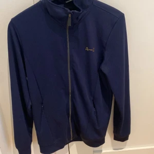 Mörkblå zipjacka från A–Z, XS - Passar dig som är 170-180 (stor k storleken). Snygg mörkblå jacka från A–Z med hel dragkedja framtill och diskret logga på bröstet. Jackan har hög krage, två fickor med dragkedja och är tillverkad i ett mjukt, stretchigt material som känns sportigt och bekvämt.