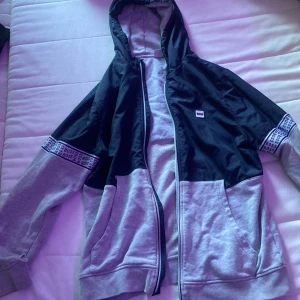 Svart och grå vindjacka från BOSS - Snygg vindjacka från BOSS i svart och grått med huva och dragkedja. Jackan har logoband på ärmarna och stora fickor framtill. Perfekt för dig som gillar sportig och clean stil. Materialmix med polyester och bomull för både komfort och stil.