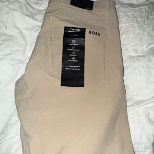 Beige byxor från BOSS med stretch - Snygga beige byxor från BOSS med 4-vägsstretch och vattenavvisande material. De är lätta, snabbtorkande och har en diskret logga på bakfickan. Perfekta för en clean och sportig look.