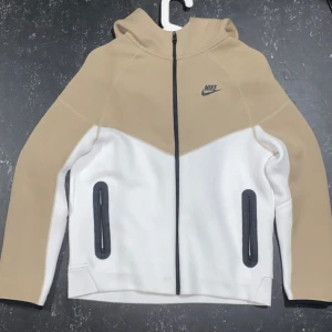 Nike tech fleece - Nike hoodiejacka i beige och vitt med svart dragkedja och svarta detaljer vid fickorna. Jackan har huva, lång ärm och sportig look. Passar dig som gillar streetwear och vill ha en snygg och bekväm jacka till vardags. Märkt med Nike-logga på bröstet.