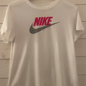 Vit Nike t-shirt med logga - Fräsch vit t-shirt från Nike med klassisk logga i rosa och grått på bröstet. Rund hals och korta ärmar, tillverkad i mjuk bomull som är skön mot huden. Perfekt för dig som gillar sportig och enkel stil med en tydlig street vibe. Den är endast använd fåtal gånger och i väldigt gott skick. Skriv gärna om det är något ni undrar eller har funderingar över😊:))