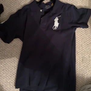 Mörkblå Polo Ralph Lauren t-shirt - Snygg mörkblå t-shirt från Polo Ralph Lauren med klassisk vit polospelare-logga på bröstet. T-shirten har korta ärmar, krage och knappar vid halsen. Perfekt för dig som gillar stilrena märkesplagg med sportig touch.