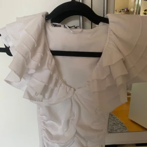 Vit volangtopp från Zara - Supersöt vit topp från Zara med flera lager volanger över axlarna och v-ringning framtill. Ärmlös modell i ribbad bomullsmix som ger en snygg passform. Perfekt för dig som vill sticka ut med en feminin och trendig look.