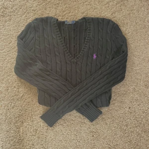 Svart stickad tröja från Polo Ralph Lauren - Svart kabelstickad tröja från Polo Ralph Lauren med lila broderad logga på bröstet. V-ringad hals och långa ärmar med ribbade muddar. Klassisk och snygg passform som funkar till jeans eller kjol. Perfekt för dig som gillar stilrena detaljer.