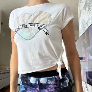 Vit croppad t-shirt med hjärta - Säljer en vit croppad t-shirt med holografiskt hjärta och texten 'Blow your heart out' på bröstet. T-shirten har korta ärmar och en snygg knytning i sidan som ger en cool vibe. Perfekt för dig som gillar unika detaljer och statement-plagg. Jag har vuxen storlek S