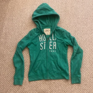 Grön hoodie från Hollister med dragkedja - Snygg grön hoodie från Hollister med vit text på bröstet. Tröjan har huva med snörning, två fickor fram och dragkedja.Mjuk bomullskänsla och långärmad modell.