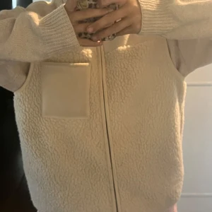 Beige fleeceväst med ficka - Mysig beige fleeceväst med dragkedja framtill och en fyrkantig bröstficka. Västen har en fluffig, teddy-liknande yta och är ärmlös med mjuka kanter. Perfekt att slänga över en hoodie eller stickad tröja för extra värme.