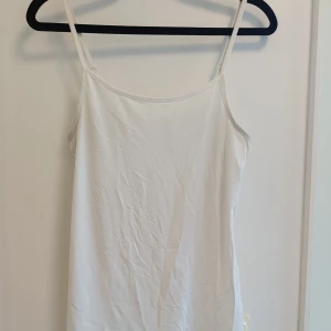 Vitt linne  - Säljer en enkel och stilren vitt linne med smala axelband och ärmlös design. Linnet har en rak passform som ger en clean look. Perfekt för varma dagar eller att styla med accessoarer för en personlig touch.