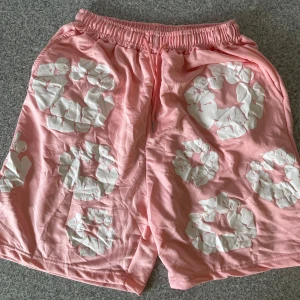 Rosa shorts från Denim Tears med tryck - Säljer ett par rosa shorts från Denim Tears med vita blommiga tryck över hela plagget. Modellen har elastisk midja med snörning och en bakficka. Materialet är mjukt och känns som bomull, perfekt för chill dagar. Unik design som sticker ut.