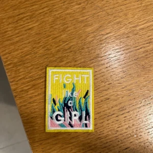 Broderat tygmärke Fight Like a Girl - Färgglatt broderat tygmärke med texten 'FIGHT like a GIRL!' på gul bakgrund och coola blå, rosa och svarta eldflammor. Perfekt att sy fast på jackan, väskan eller jeansen för en extra statement-look.