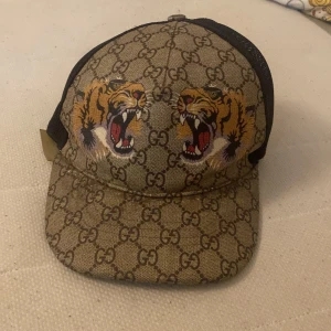 Gucci keps med tigertryck och GG-mönster - Snygg keps från Gucci med klassiskt GG-monogram i beige och brunt, prydd med två coola tigerhuvuden framtill. Baksidan är i svart mesh och justerbar med rem i grönt och rött. Perfekt statement-piece för dig som vill sticka ut.