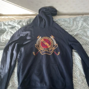 Marinblå hoodie från U.S. Polo Assn - Snygg marinblå hoodie från U.S. Polo Assn med stort broderat emblem i rött, gult och beige på bröstet. Tröjan har huva, känguruficka och långa ärmar. Perfekt för dig som gillar sportig och klassisk stil.