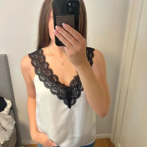 Vit topp med svart spets - Supersnygg vit linnetopp med breda svarta spetsdetaljer längs v-ringningen och axelbanden. Sidenliknande material som känns lyxigt och mjukt mot huden. Aldrig använd💕