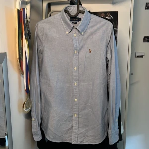 Skjorta från Ralph Lauren  - Klassisk ljusblå skjorta från Ralph Lauren i bomull med broderad logga på bröstet. Skjortan har lång ärm, knappar framtill och button down-krage. Snygg custom fit som sitter riktigt bra och passar till många olika stilar.