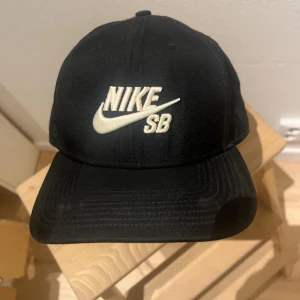 Svart Nike SB keps med broderad logga - Svart keps från Nike SB med broderad vit logga framtill. Klassisk böjd skärm och ventilerande mesh på insidan för extra komfort. Perfekt för skate eller streetwear. Justerbar passform och stilren design som funkar till det mesta.