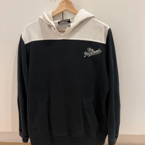 Svart och vit hoodie från The Hundreds - Snygg hoodie från The Hundreds i svart och vitt med broderad logga på bröstet. Tröjan har huva med metallöglor, känguruficka och långärmade ärmar. Perfekt för dig som gillar streetwear och vill ha en chill look.