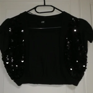 Svart bolerotopp från H&M - Snygg svart bolerotopp från H&M med glittriga paljetter på framsidan. Perfekt för att lägga till lite glamour till din outfit. Kortärmad design som passar bra till festliga tillfällen.