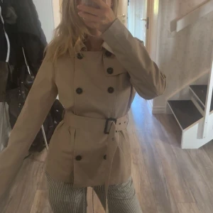 Beige trenchcoat med bälte - Snygg beige trenchcoat från Jacqueline de Yong med klassisk krage, dubbelknäppning och bälte i midjan. Jackan har långa ärmar och två fickor framtill. Perfekt till ett par jeans eller kostymbyxor, till en utekväll eller bara en vanlig sval höstdag.