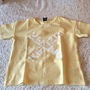 Gul Kenzo t-shirt med vit logga - Kenzo t-shirt i ljusgul färg med vit, stor logotyp tryckt diagonalt över bröstet. Klassisk rund hals och korta ärmar. Tillverkad i mjuk bomull som känns skön mot huden. Perfekt för dig som gillar stilrena och trendiga märkesplagg.