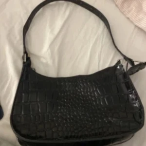 Svart axelväska i croco-imitation - Snygg svart axelväska med croco-mönstrad yta och dragkedja upptill. Väskan har en stilren, avlång form och ett bredare axelband. Perfekt storlek för det viktigaste och ger en trendig vibe till din outfit.