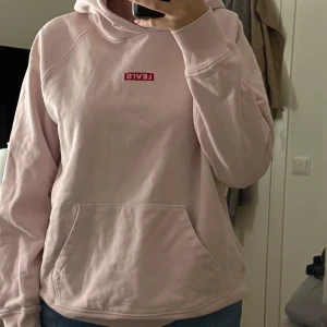 Ljusrosa hoodie från Levi's - Säljer en ljusrosa hoodie från Levi's i storlek M. Tröjan har en klassisk magficka, huva och Levi's-logga i rött på bröstet. Perfekt för chill dagar och enkel att matcha med jeans eller leggings. Knappt använd.