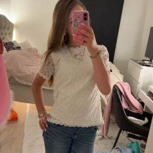 Vit spetsblus med kort ärm - Säljer en söt vit blus i spets med korta ärmar och rund hals. Blusen har fina spetsdetaljer över hela och vågig kant nertill. Perfekt att styla med jeans eller kjol för en fräsch look. Använt typ 5 gånger. Storlek xs från veromoda.