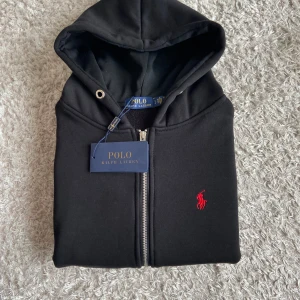 Svart hoodiejacka från Polo Ralph Lauren - Svart hoodiejacka från Polo Ralph Lauren med dragkedja och huva. Klassisk röd broderad logga på bröstet och kängurufickor framtill. Tillverkad i mjuk bomull för en skön och avslappnad stil. Perfekt för dig som gillar stilrena och sportiga plagg.
