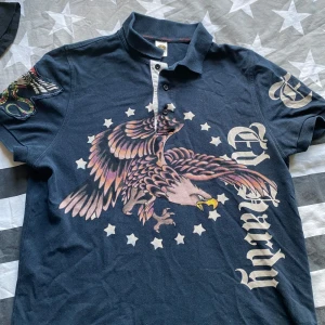 Ed Hardy navy t-shirt med örntryck - Mörkblå Ed Hardy t-shirt med stort örntryck och stjärnor på framsidan, samt logga på både fram- och baksida. Klassisk krage med knappar och coolt tryck på ena ärmen. Materialet är mjuk bomull och passformen är normal.