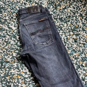 Mörkblå skinny jeans från Nudie - Säljer ett par mörkblå skinny jeans från Nudie Jeans i modellen Tight Long John. Klassisk femficksdesign, snygga kontrastsömmar och Nudie-logga på bakfickan. Jeansen är i stretchig denim som sitter tight hela vägen ner.