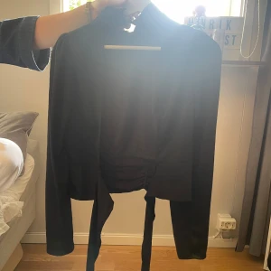 Svart och beige omlottblus med knyt - Två blusar i svart och beige med lång ärm, puffade axlar och snygg omlottknytning baktill. Båda har rynkade detaljer vid midjan och knytband som ger en cool siluett. Perfekt för dig som gillar trendiga och unika plagg med lite extra attityd.