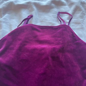 Lila sammetstopp med smala axelband - Säljer en snygg lila topp i mjuk sammet med smala axelband. Toppen har en enkel, figurnära passform och är perfekt för dig som vill sticka ut med en cool färg och lyxig känsla. Passar till både jeans och kjol för en trendig look.