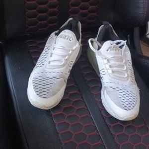 Nike Air Max 270 vita sneakers - Nike Air Max 270 sneakers i vitt med meshpaneler och svarta detaljer. Snygg design med stor synlig Air-enhet i hälen och klassisk Nike-logga på sidan. Skorna har snörning och en sportig vibe, perfekta för dig som gillar streetstyle.