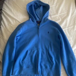 Blå hoodie från Polo Ralph Lauren - Snygg blå hoodie från Polo Ralph Lauren med dragkedja och huva. Tröjan har två fickor framtill och det klassiska Polo-logot broderat på bröstet. En oanvänd polo- Ralph lauren tröja som är i topp skick🌟