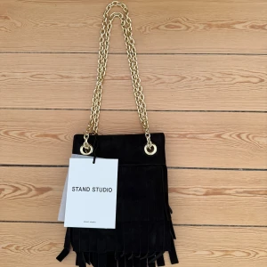 Svart mocka axelväska från Stand Studio - Svart axelväska i mocka från Stand Studio med fransdetaljer och guldig kedja som axelrem men kan bäras längre som cross body. Väskan har en fyrkantig form och cool boho-vibe, perfekt för att lyfta din outfit. Modellen heter Rhea Fringe Bag. 