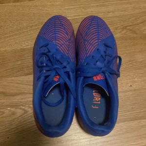 Blå Adidas Predator fotbollsskor - Säljer ett par blå Adidas Predator fotbollsskor med röda detaljer och mönster. Skorna har snörning och är tillverkade i syntetmaterial för bästa grepp och känsla på planen. Perfekta för dig som vill sticka ut med stilrena och sportiga skor. Säljs för att de är förr små för mig nu. Priset kan sänkas.
