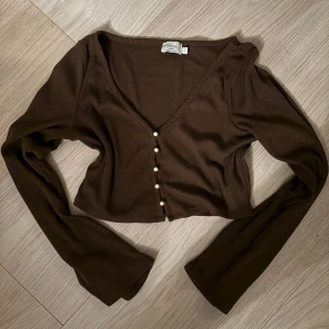 Brun croppad ribbad topp med pärlknappar - Säljer en brun croppad topp från nakd med långa ärmar och v-ringning. Toppen är ribbad och har snygga pärlknappar framtill som detalj. Perfekt att styla med höga jeans eller kjol för en trendig look.