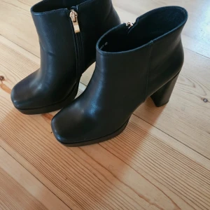 Svarta stövletter med klack och dragkedja - Snygga svarta stövletter i skinnimitation med chunky hög klack och platåsula. Skorna har rund tå och guldfärgad dragkedja på sidan för enkel påtagning. Perfekta för dig som vill ha en stilren och edgy look.